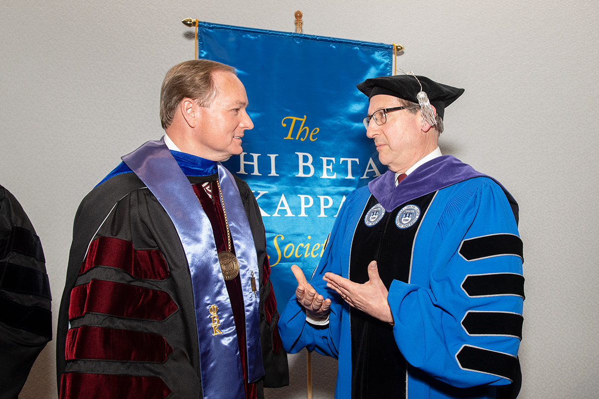 Man in black graduate robes with blue stole speaking with man in blue and black regalia in front of blue Phi Beta Kappa banner