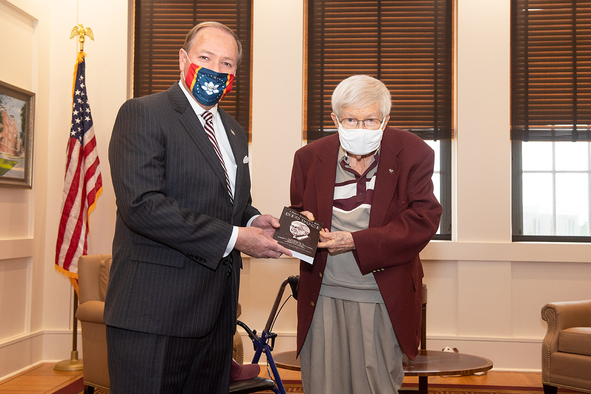 Man in dark suit and mask being presented a book by man in maroon jacket and mask