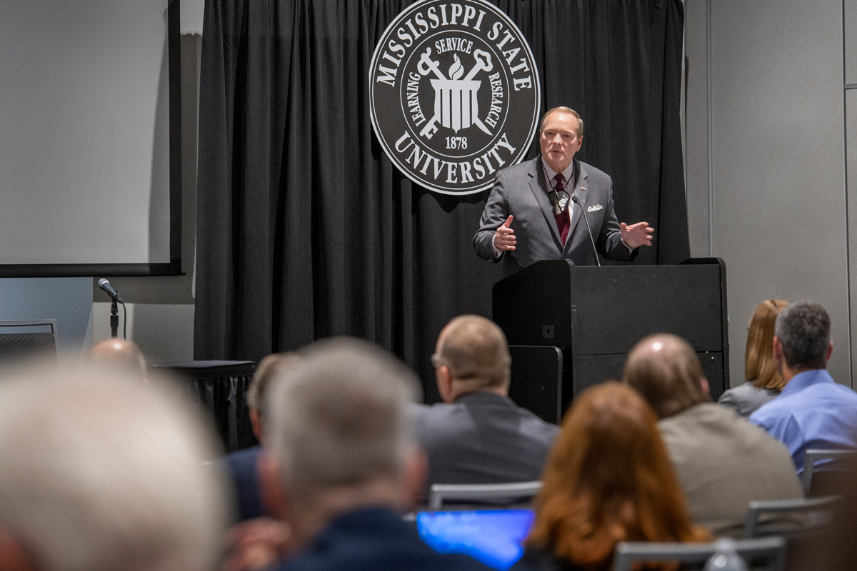 President Keenum speaks in from a podium with a black pipe and drape and MSU seal behind, with attendees heads in the foreground