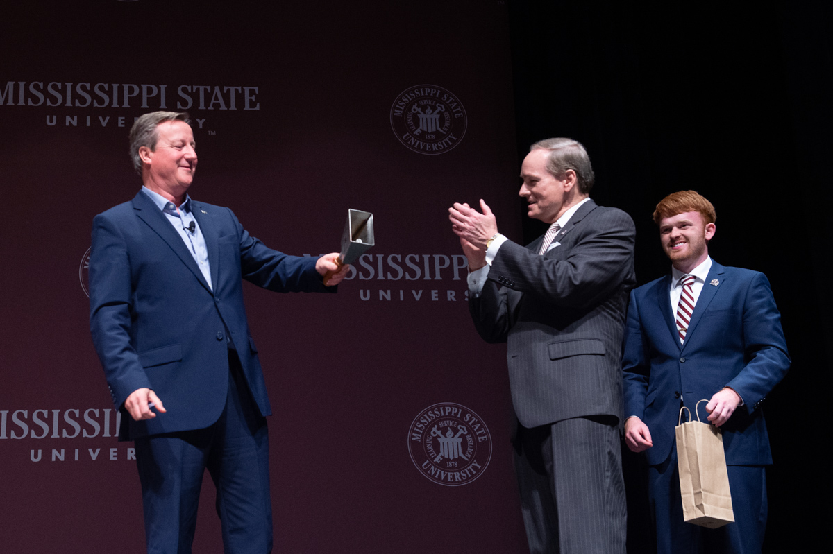 Former UK Prime Minister, David Cameron (at left), rings a cowbell while President Keenum and SA President Guest clap and smile.