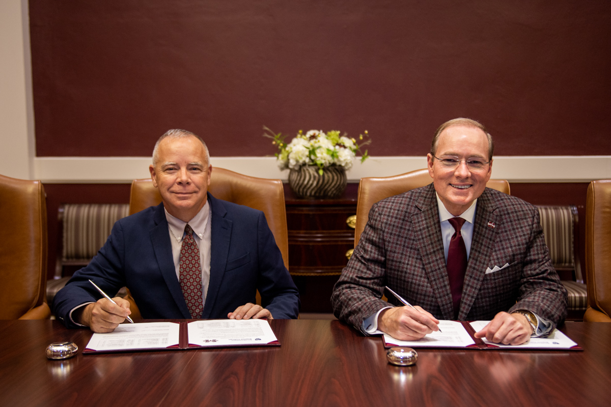 Two male university presidents each sit at a large conference style table with pens in hand