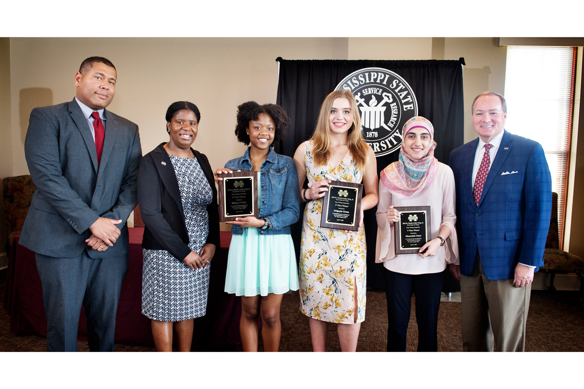 Three high school Martin Luther King Jr. Essay Writing Contest winners award winners stand between MSU VIPs, holding plaques.
