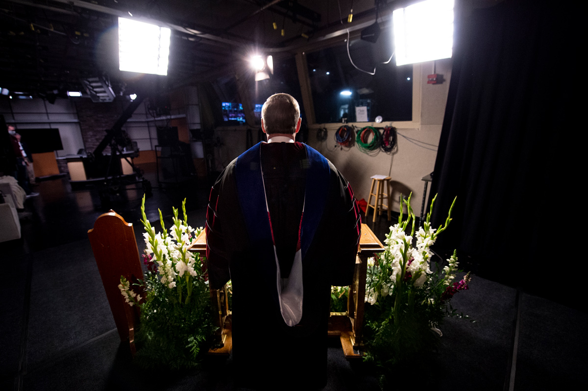 In full regalia and surrounded by podium flowers in the TV studio, President Keenum speaks towards the video cameras and lights.