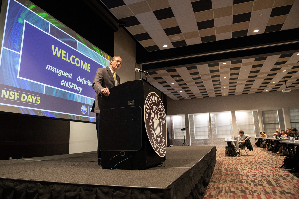 President Keenum speaks from a platform in the Mill's largest ballroom, with a slide welcoming everyone to NSF Days. 