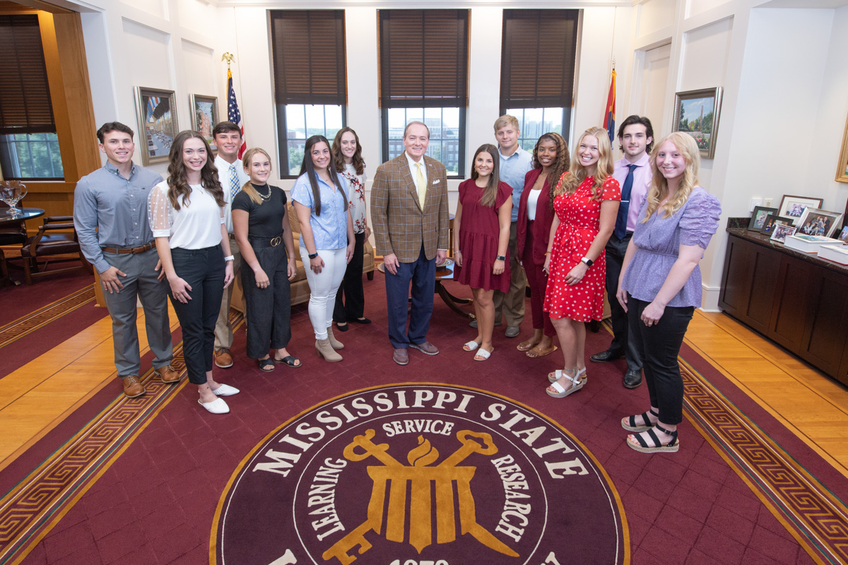 A group of 12 Presdiential Partnership Scholars pose with Keenum at the MSU seal in the Presidential Suite.
