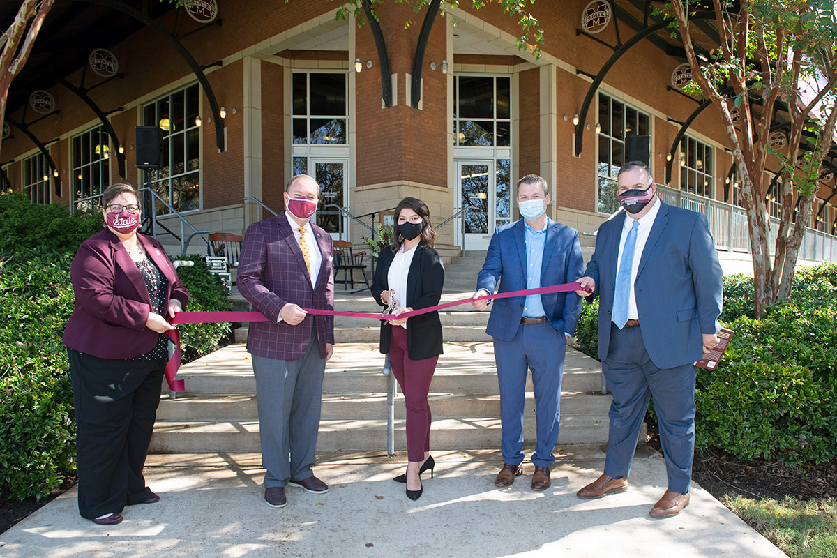 Two women and three men pose for ribbon cutting photo in front of building