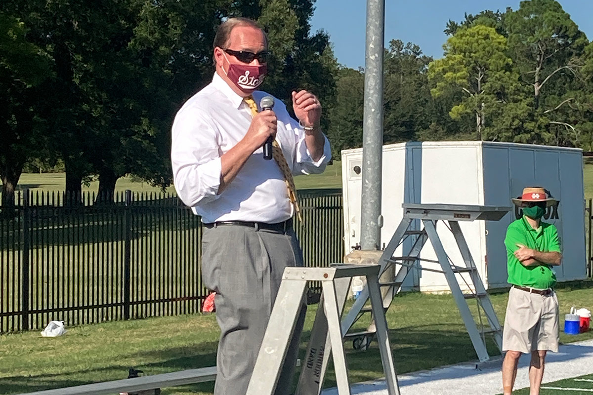 Man in white dress shirt and maroon mask speaking in microphone from ladder
