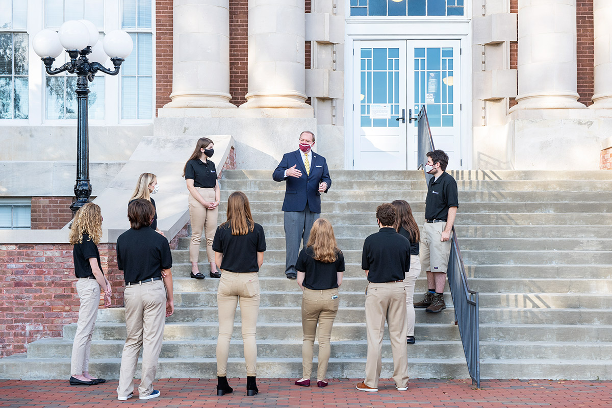 Man in maroon mask and blue blazer on steps speaking to students in masks and black polos with khakis.