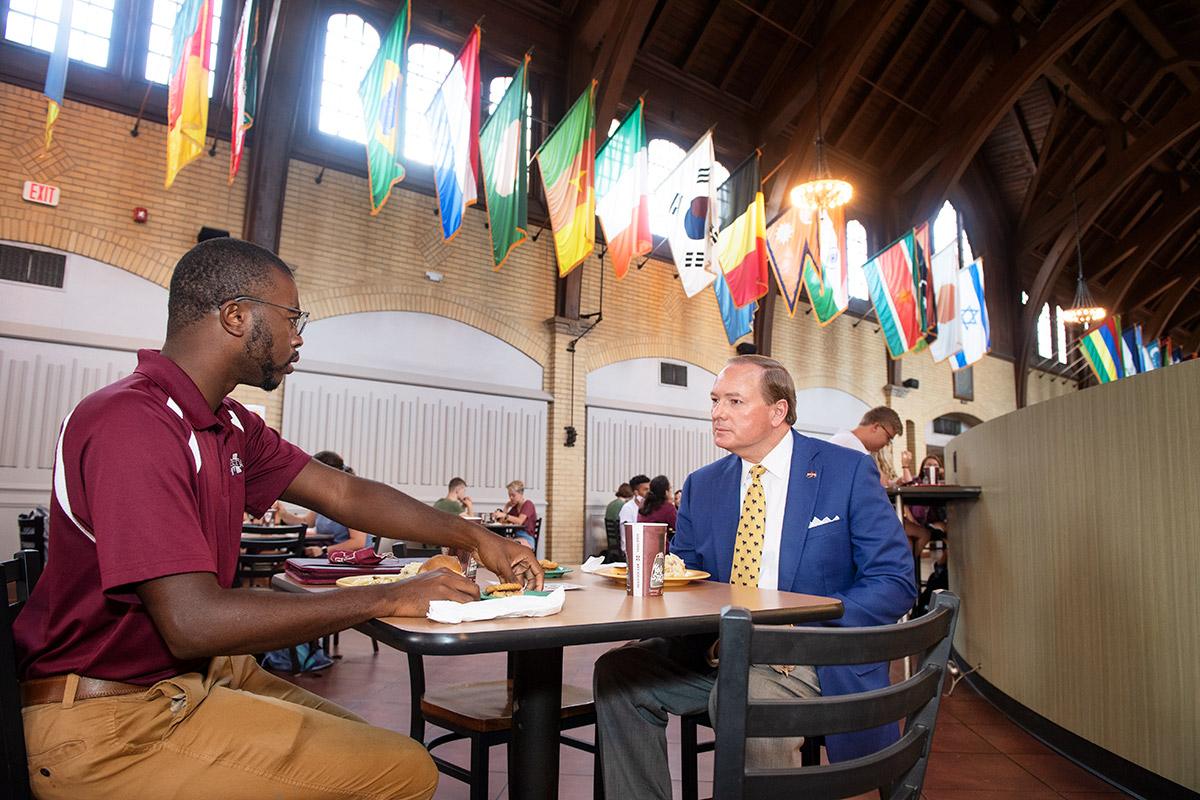 Guy in maroon polo and man in blue suit jacket talking over lunch with international flags in background