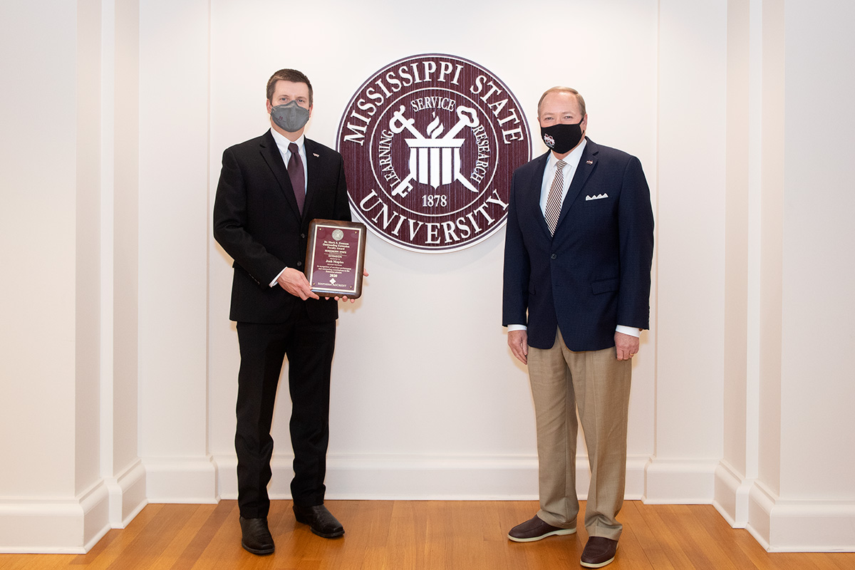 Two men in suits and face coverings in front of MSU seal. Man on the left is holding a maroon plaque.