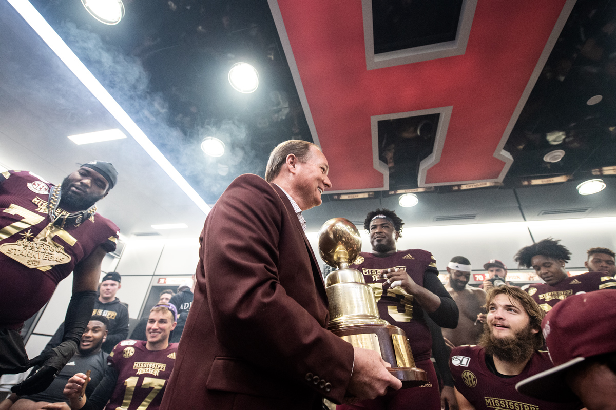 President Keenum holds the Egg Bowl in the Davis Wade locker room, while congratulating the smiling players encircling him.