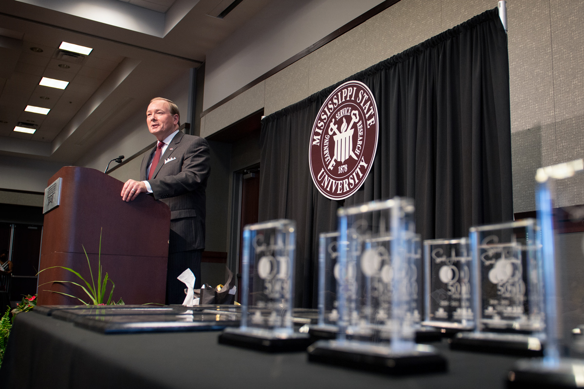 President Keenum speaks from a podium, with glass awards in the foreground on a table.