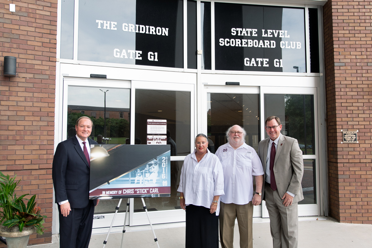 Standing in front of the glass doors to Gate G1 of the football stadium with a sign honoring Chris "Stick" Carl.
