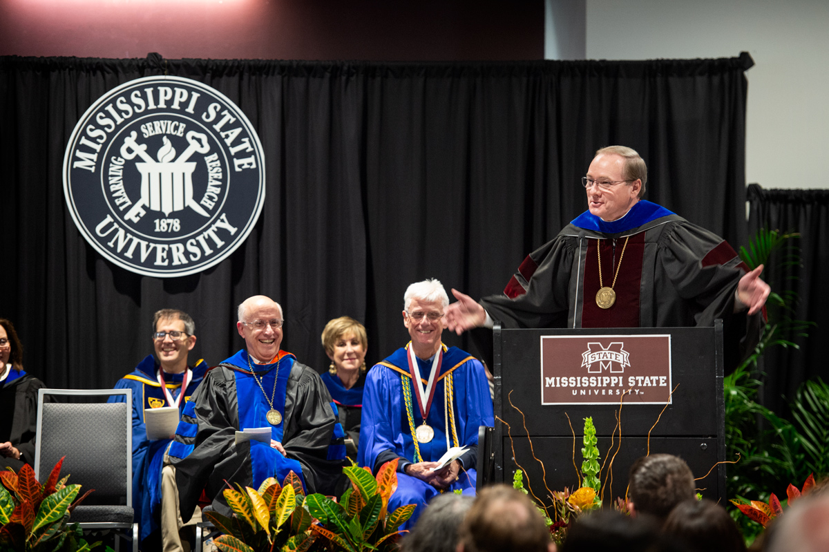 President Keenum speaks from the podium with his arms outstretched while fellow administrators listen in full regalia.