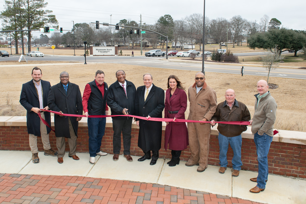 The group of VIP ribbon-cutters pose with maroon ribbon with the intersection of Blackjack Road and Hailstate Boulevard behind.