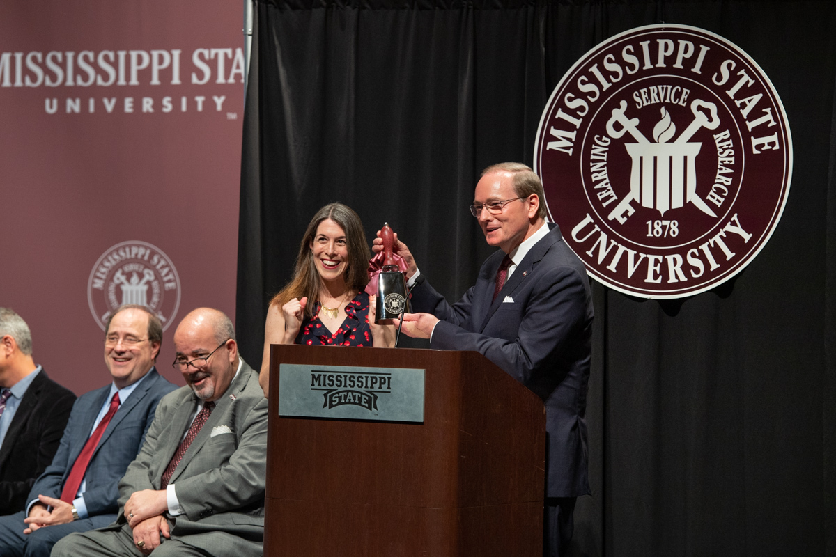 Poet Laureate Catherine Pierce eceives an engraved cowbell from President Keenum, with the MSU seal behind the podium.