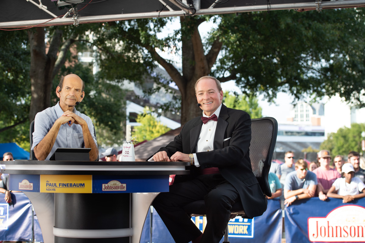 Paul Finebaum and President Keenum smile from  the table on the set of the on location studio, with the Junction behind them.