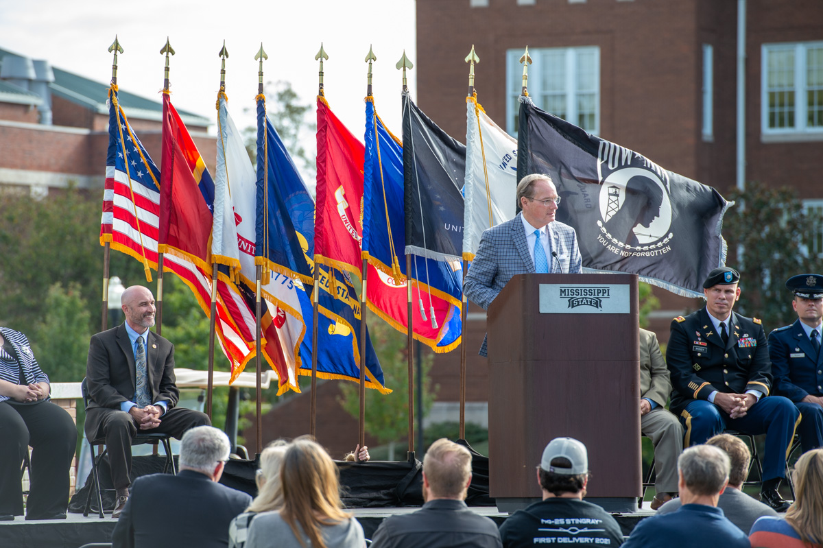 President Keenum speaks from a podium on the Drill Field with flags blowing in the breeze behind him.