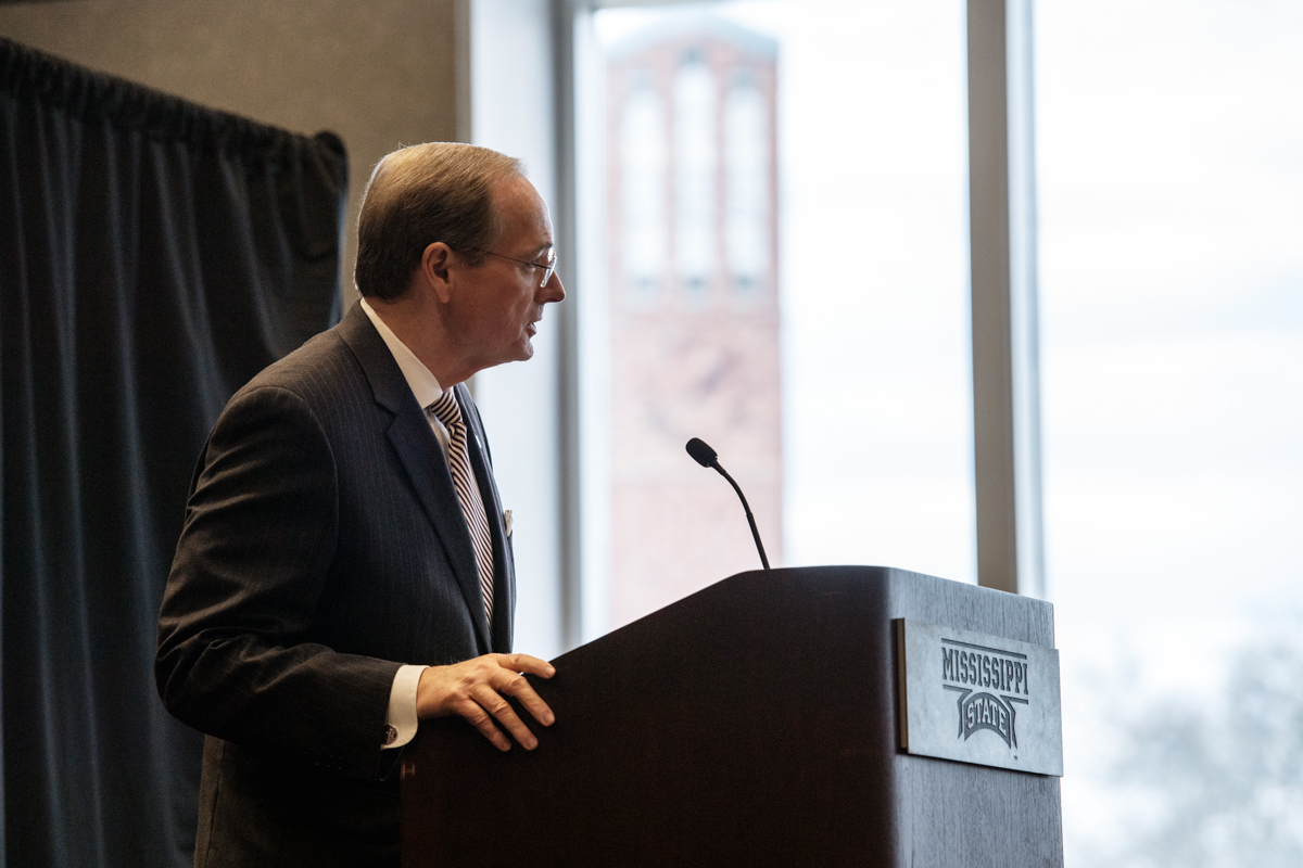 A profile angle on President Keenum speaking at the podium, with the Chapel of Memories tower visible through the window beyond.