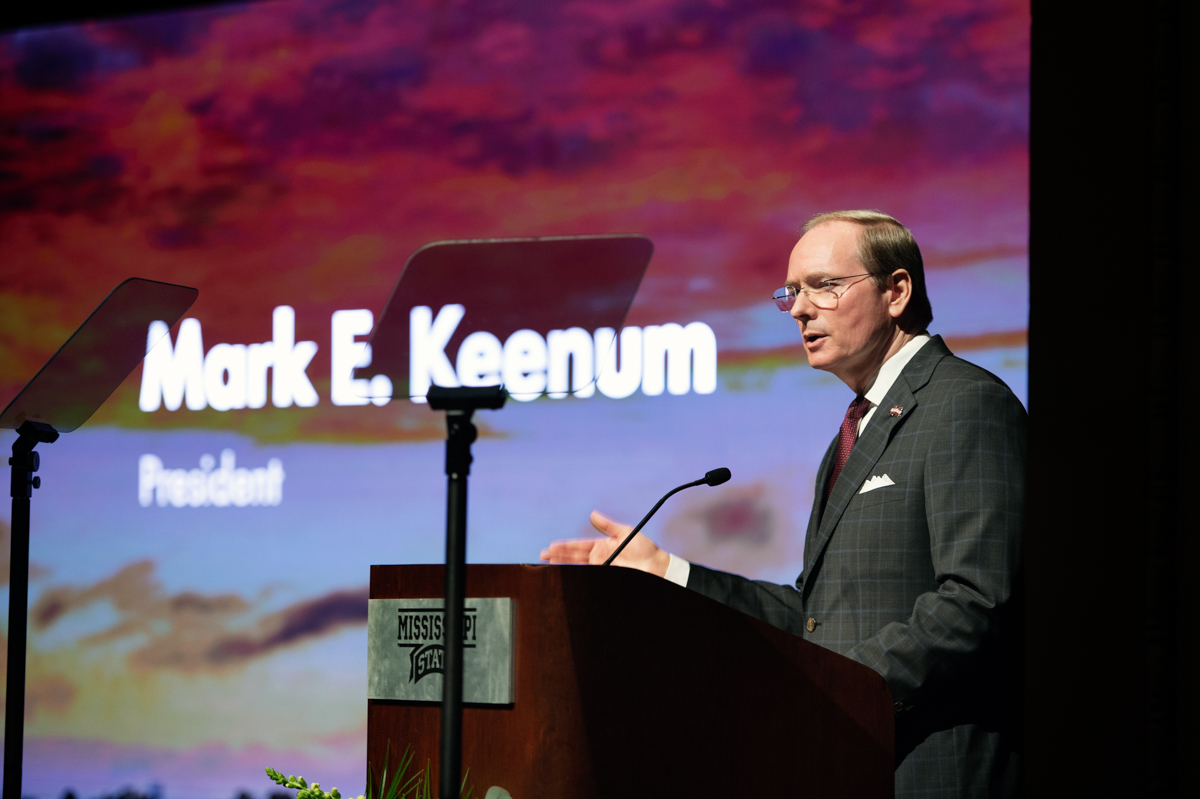 President Keenum speaks from the podium, with his name projected onto a firey sunset image on the screen behind.