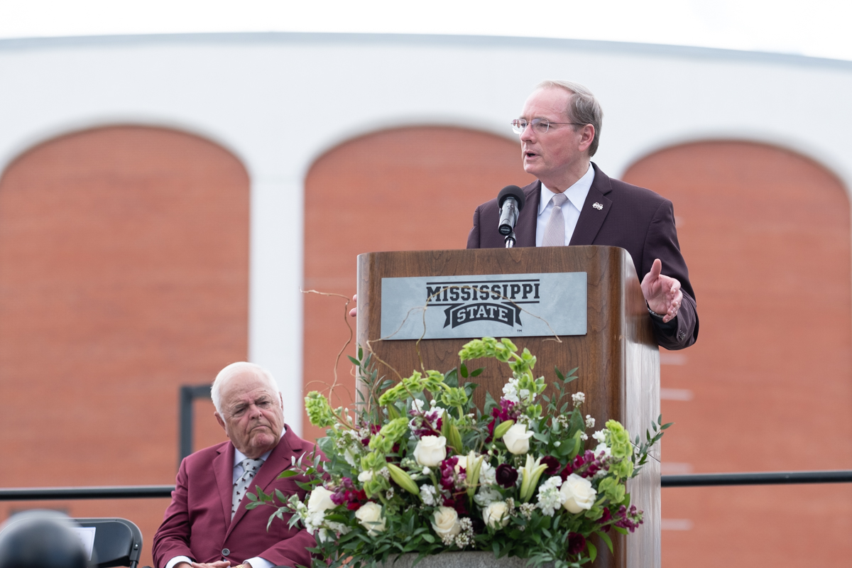 President Keenum speaks from an outdoor podium with The Hump as a backdrop and Coach Polk listening from his seat on the stage.